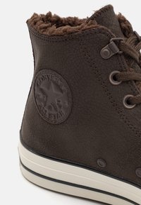 Hnědá kožená vysoká teniska s texturovaným povrchem, umělou kožešinou uvnitř, kulatou nášivkou loga a bílou gumovou podrážkou.