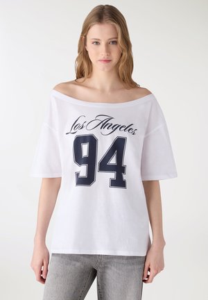 Giovane donna che indossa una maglietta bianca larga con scollo a barca con la scritta "Los Angeles" e il numero 94 grande, abbinata a jeans grigio chiaro, in piedi contro uno sfondo semplice.