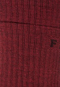 Calcetín de lana roja con textura acanalada, que presenta un pequeño logotipo negro cerca de la parte superior. El tejido parece suave y cálido.