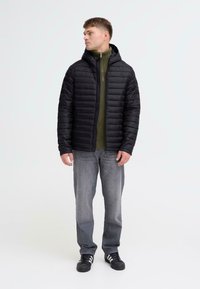 Sort polstret jakke med hætte, lynlåslukning og struktureret overflade, over en grøn højhalset top, parret med grå jeans og sorte sko.