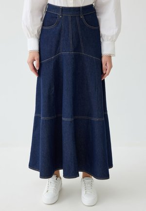Femme portant une longue jupe en denim bleu foncé avec des coutures visibles, des baskets blanches et un chemisier blanc à manches longues avec des poignets en dentelle.