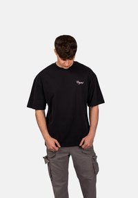 Černé bavlněné tričko s uvolněným střihem, krátkými rukávy a malým bílým vyšívaným logem "Crystal" na levé části hrudi.