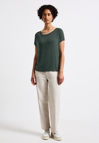 Top corto a maniche corte di un verde scuro, in tessuto leggero, con scollatura rotonda, abbinato a pantaloni ampi di un beige chiaro e sneakers bianche.