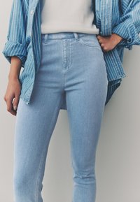 Blugi bleu deschis cu talie înaltă și croială mulată, asortați cu o cămașă albastră cu dungi, largă, cu nasturi. Textură de denim netedă, detalii minime.