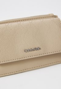 Beige lommebok laget av teksturert materiale med et preget sirkulært mønster og en sølvfarget Calvin Klein-logo på forsiden.