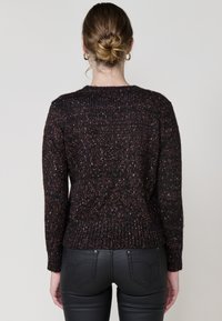 Dunkelbrauner Strickpullover mit mehrfarbigen Sprenkeln, ausgestattet mit einem gerippten Ausschnitt und Bündchen, kombiniert mit schwarzen, engen Hosen.