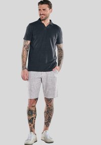 Dunkelgraues Poloshirt, weiße Shorts mit Streifen und weiße Turnschuhe. Kurzärmlig, Standardkragen und sichtbare Tattoos an Armen und Beinen.