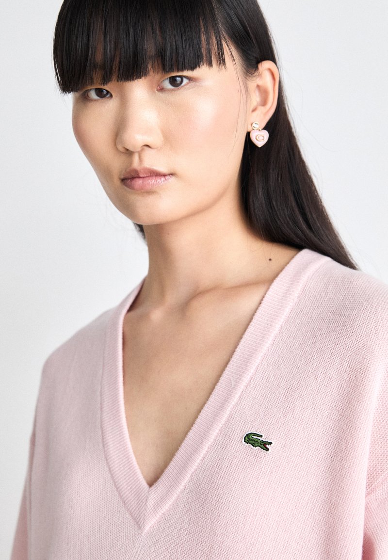 Pull V en maille douce rose clair avec un petit logo de crocodile vert sur la poitrine. Le modèle porte des boucles d'oreilles en forme de cœur avec une finition brillante.