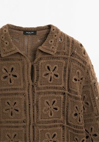 Cardigan en crochet marron avec motif floral, présentant un col, une fermeture à boutons et un tissu texturé.
