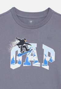 Szürke póló, amelyen egy hódeszkás grafikája látható a "GAP" felirat fölött. A dizájn kék és fehér hegyi tájat és fákat ábrázol.