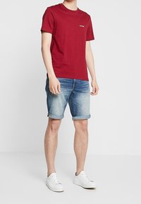 Röd bomull t-shirt med logotyp på bröstet, som matchas med ljusblå denimshorts och vita sneakers. Slät textur genom hela plagget.