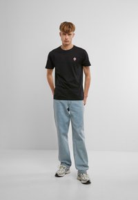 Sort kortærmet t-shirt med et pink donutsdesign på venstre bryst, parret med lyseblå lige ben jeans og sportssko.