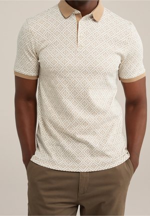 Mann trägt beige Poloshirt mit beigem geometrischem Muster und Kragen, kombiniert mit brauner Hose, Hände in den Taschen, neutraler Hintergrund.