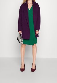 Lila übergroßer Mantel, grünes Wickelkleid, burgunderfarbene gehäkelte Absätze und eine gemusterte schwarz-weiße Clutch. Glatte Stofftexturen.