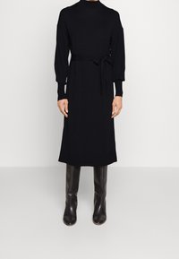 Robe noire à manches longues avec un col haut, une taille ceinturée et des poignets côtelés. Portée avec des bottes sombres hautes jusqu'aux genoux sur un fond uni.