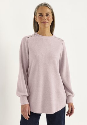Moden kvinde med gråt hår iført en lyserød ribstrikket sweater med knapdetaljer på skuldrene og mørke bukser mod en ensfarvet baggrund.