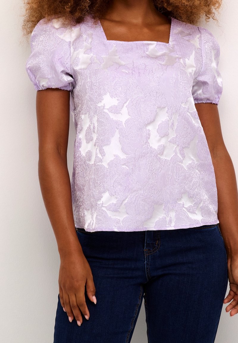 Donna che indossa un top floreale in jacquard lilla con maniche a palloncino e scollo quadrato, abbinato a jeans blu scuro.