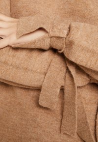 Gros plan sur des bras croisés portant un pull en tricot beige avec des poignets à trou pour le pouce et une ceinture assortie nouée à la taille.