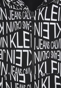 Felpa nera con cappuccio e testo bianco, caratterizzata dal nome del marchio ripetuto "CALVIN KLEIN JEANS." Il materiale è liscio, con zip anteriore e tasche frontali.