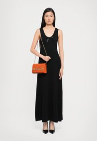 Femeie îmbrăcată într-o rochie lungă neagră cu o fundiță mică, cu tocuri negre și ținând o geantă de umăr Love Moschino portocalie cu un lanț.
