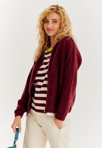 Burgundské pletené cardigan s voľným strihom, otvoreným predným dielom a dlhými rukávmi, prehodený cez pruhované tričko. Mäkko štruktúrovaný a ležérny dizajn.