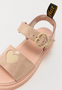 Roza bleščične sandale s srčasto izrezano obliko in zaponko na trak. Podrobnost mehkega notranjega podplata, teksturirana zunanjost in zanka za poteg.