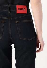 Jeans in denim blu scuro con cuciture arancioni, dotati di una etichetta rossa "HUGO" sul retro in vita e due tasche posteriori.
