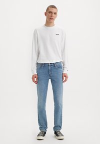 Jonge man die staat in een wit Levi's langlijfshirt, lichtblauwe spijkerbroek en zwarte Converse-achtige sneakers op een witte achtergrond.