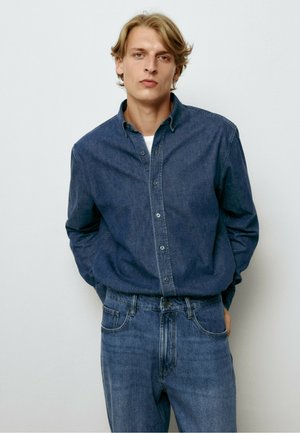 Ung mand med blondt hår iført en mørkeblå denimskjorte over en hvid undertrøje og blå jeans, stående foran en ensfarvet hvid baggrund.