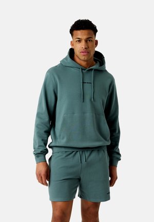 Mees kannab türkiissinist kapuutsiga pusa ja sobivaid lühikesi pükse, seisab ühevärvse heleda tausta ees ja vaatab otse eespool neutraalse ilmega.