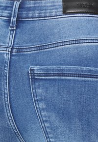 Jeans de denim azul com cintura alta, apresentando um bolso traseiro e uma pequena etiqueta de marca em couro preto perto da cintura. Textura suave.