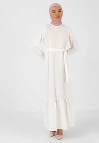 Long white dress with long sleeves, a ruffled hem, and a white belt. Worn with a light pink headscarf and nude high heels.