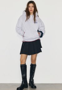 Grijze hoodie met voorzak, gecombineerd met een zwarte geplooide rok. Zwarte kniehoge laarzen met gespen. Laagjes met rode en blauwe rand.