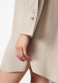 Beige Hemdkleid mit langen Ärmeln, das zwei Knöpfe an den Manschetten hat. Der Stoff hat eine glatte Textur und eine leicht lockere Passform.