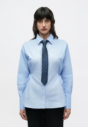 Svetlomodrá košeľa na gombíky s výrazným golierom, dlhými rukávmi a bočnými zaväzovacími detailmi. Doplnok tvorí tmavomodrá pruhovaná kravata. Minimalistický dizajn.