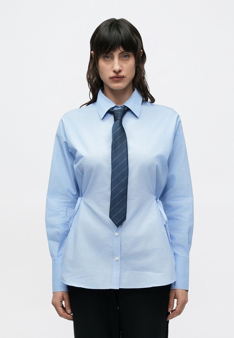 Ανοιχτό μπλε πουκάμισο με κουμπιά, με στιβαρό γιακά, μακριά μανίκια και δεσίματα στο πλάι. Συνδυάζεται με μια σκούρα μπλε γραβάτα με ρίγες. Μινιμαλιστικός σχεδιασμός.