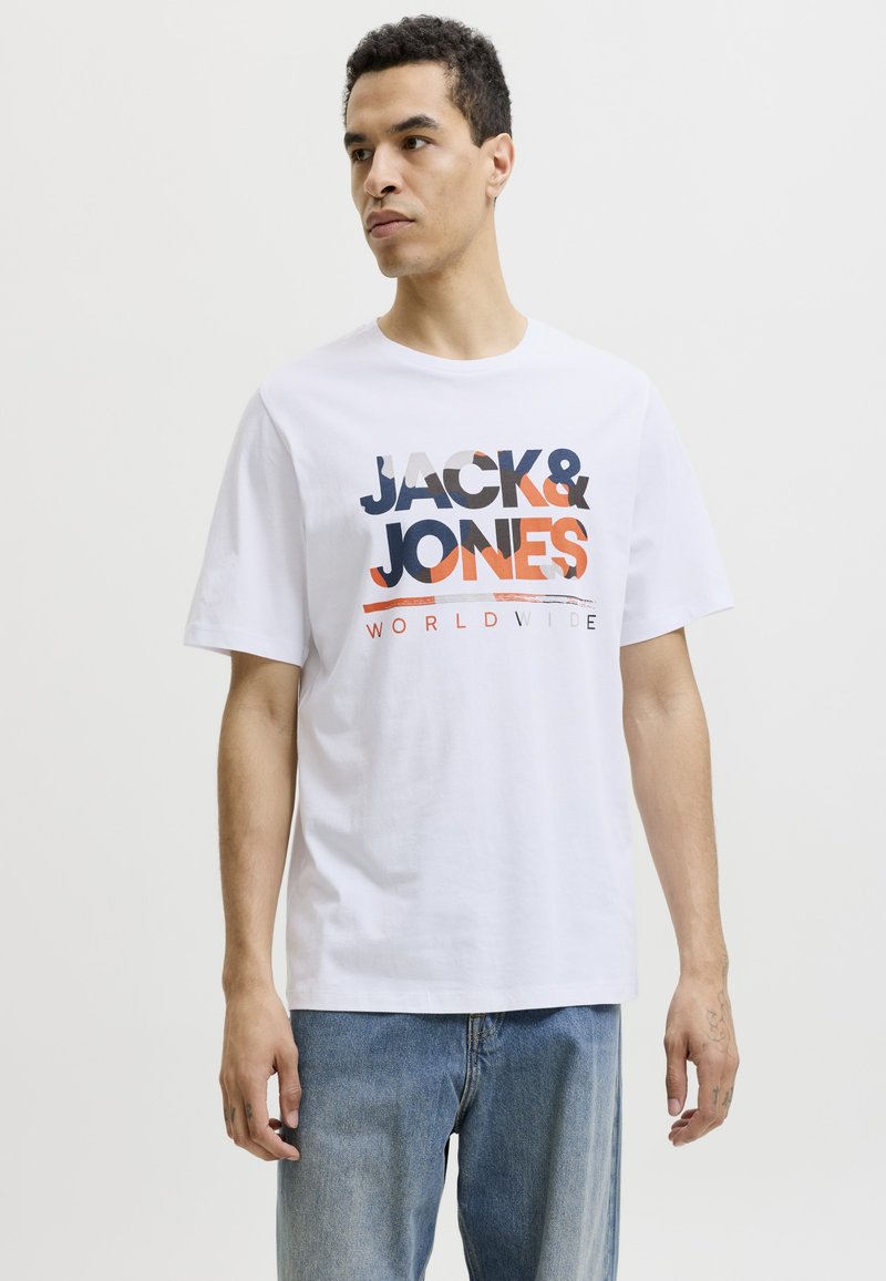 Hombre con camiseta blanca de manga corta con texto "JACK & JONES WORLDWIDE" y vaqueros azules, de pie frente a un fondo claro y sencillo.