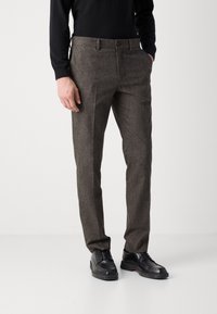 Pantaloni marroni texture per uomo con design aderente, dotati di chiusura a un bottone e tasche laterali, abbinati a scarpe di pelle nere.