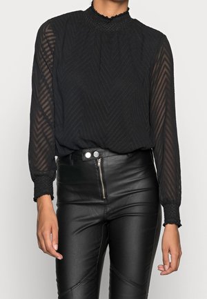 Woman wearing a black sheer long-sleeve blouse with chevron patterns and high-waist black leather pants with front zipper and snap buttons.