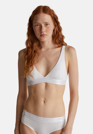 Donna dai capelli rossi con lentiggini che indossa un reggiseno bianco morbido e uno slip abbinato con elastici "United Colors of Benetton".