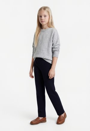 Šviesiaplaukė mergina stovi, dėvi šviesiai pilką megztinį, juodas kelnes ir rudus mokasinus, prieš vientisą baltą foną.