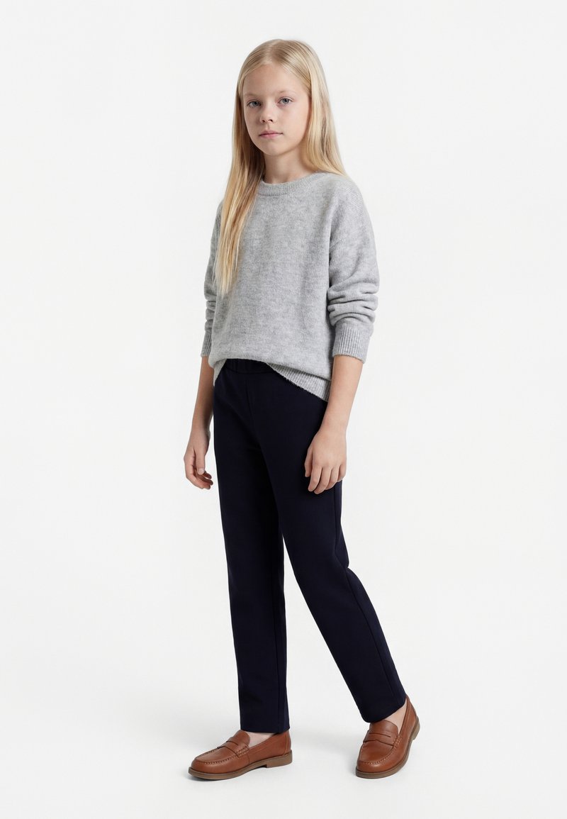 Fată blondă stând, purtând un pulover gri deschis, pantaloni negri și mocasini maro, pe un fundal alb simplu.