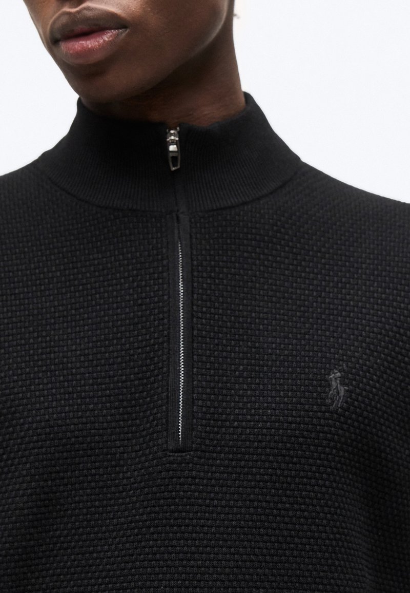Homme portant un pull noir texturé à col montant avec une demi-fermeture éclair et un petit logo brodé sur le côté gauche de la poitrine.