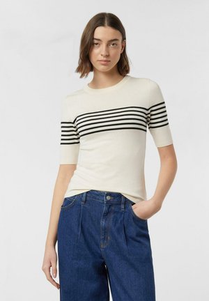 Frau trägt ein weißes kurzärmeliges Oberteil mit schwarzen horizontalen Streifen über Brust und Ärmeln, kombiniert mit hochtaillierten blauen Jeans.