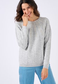 Suéter de punto gris con un cuello de canalé, patrones de punto detallados y sutiles acentos dorados, combinado con unos jeans azul claro.