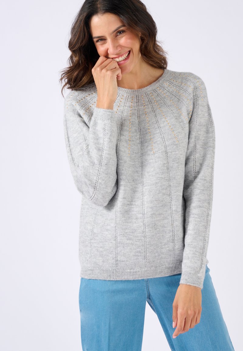 Suéter de punto gris con un cuello de canalé, patrones de punto detallados y sutiles acentos dorados, combinado con unos jeans azul claro.