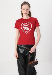 Camiseta de algodón roja con un diseño de corazón blanco que incluye el texto "Coach" y un caballo alado, combinada con pantalones de cuero negros y un bolso marrón.