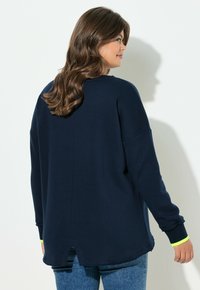 Frau mit langen braunen Haaren, die ein marineblaues Langarmshirt und blaue Jeans trägt, steht vor einer weißen Wand, hinter ihr wirft sie einen Schatten.
