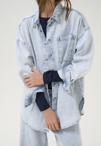 Ljusa blå oversized denimskjorta med knäppning fram, bröstficka och uppvikta ärmar. Bärs över en marinblå långärmad topp.