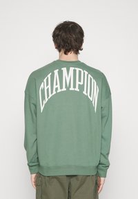 Zelená bavlněná mikina se vzorem "CHAMPION" vytištěným velkými bílými písmeny na zádech. Uvolněný střih s manžetami a lemem s žebrováním.